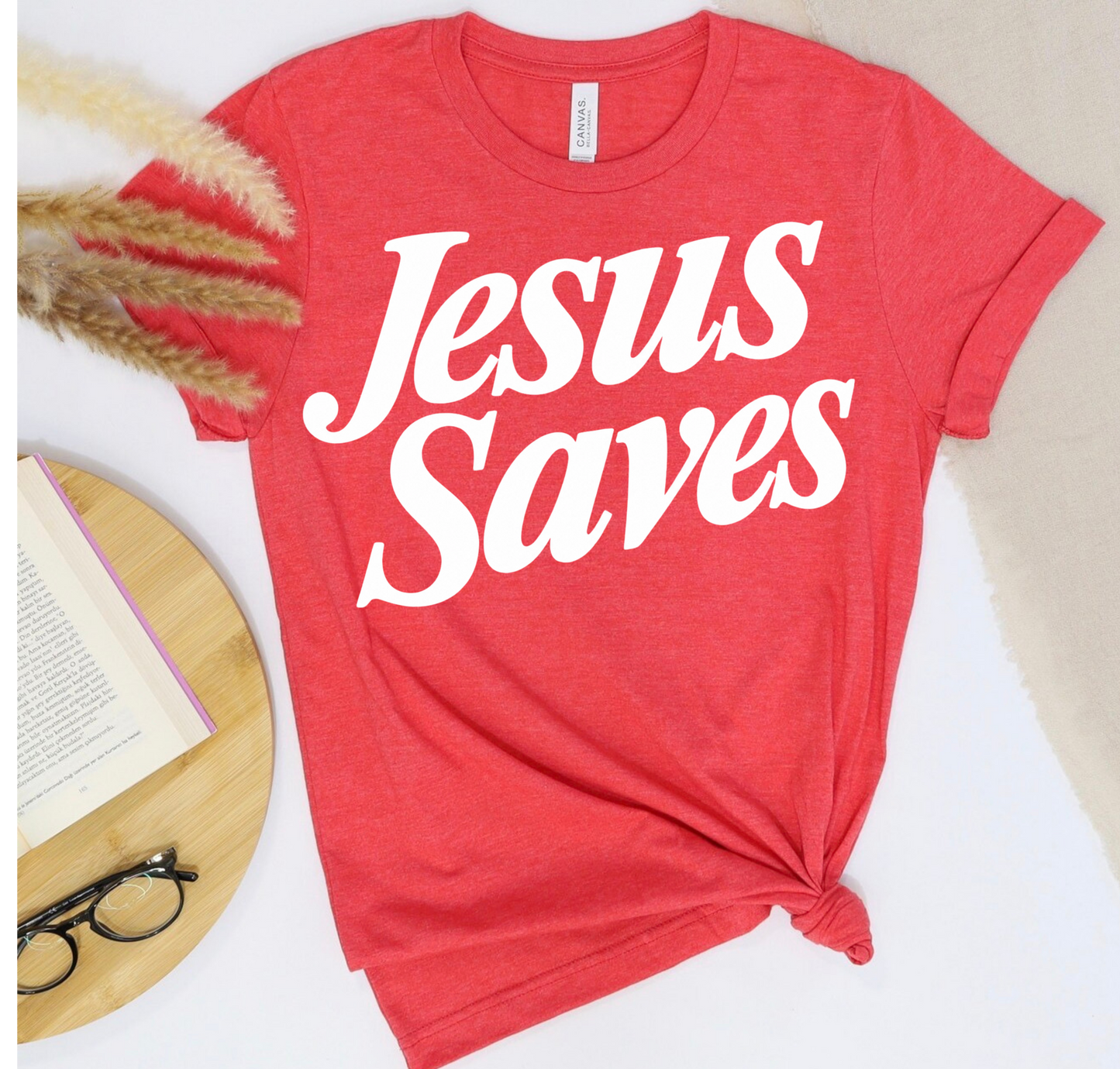 A red short-sleeve T-shirt with the phrase 'Jesus Saves' printed in large white script across the chest, styled in a flat lay with a knot at the hem. The shirt is shown beside a book, eyeglasses, dried grass, and a light neutral fabric background.